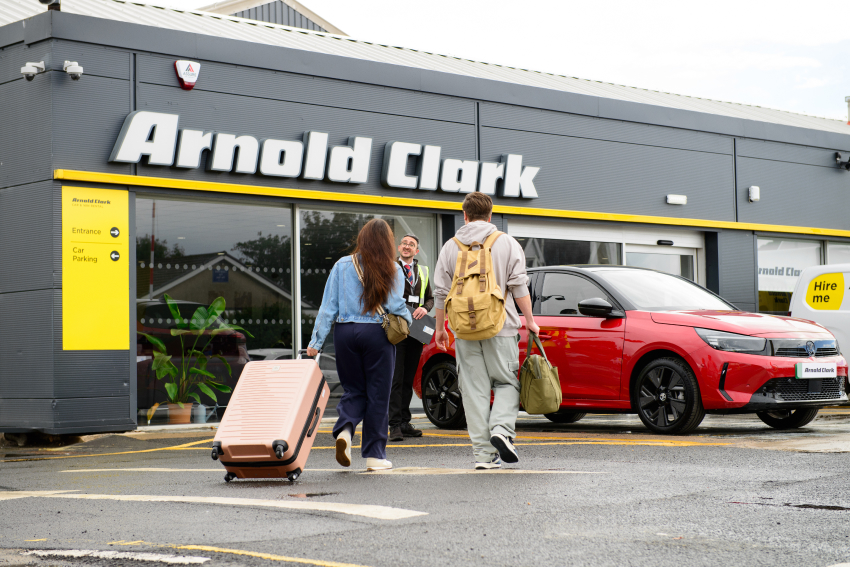 Couple with suitcases walking towards Arnold Clark Rental branch and being greeted by staff member